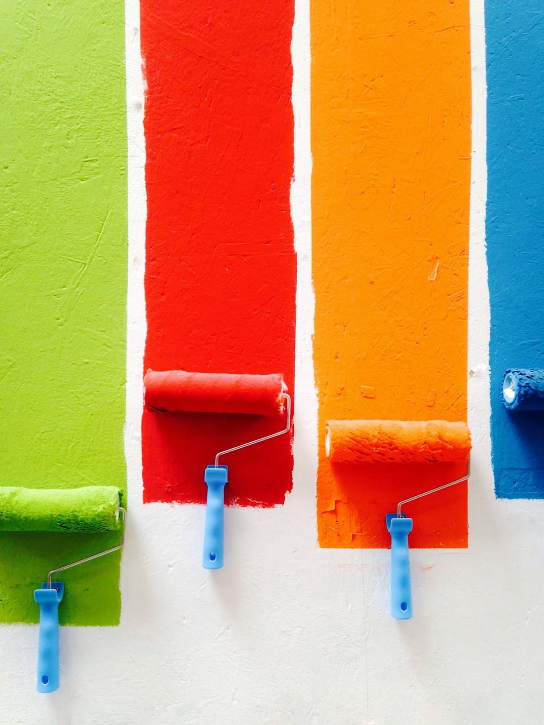 Orange, green, blue, and red paint rollers lined up on a wall